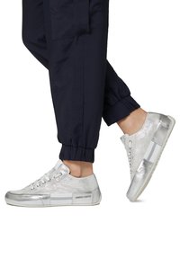 Zapatillas plateadas con un acabado metálico texturizado, cordones blancos y rayas laterales con el nombre de la marca, combinadas con pantalones informales azul marino.