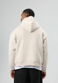 Beige hoodie met een losse pasvorm, geribbelde manchetten en onderkant, zachte textuur en een kangoeroezak, gedragen over een wit T-shirt.
