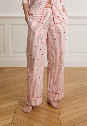 Femme portant un pantalon de pyjama blanc à rayures verticales rouges et petits cœurs rouges éparpillés, debout pieds nus sur un sol en bois.