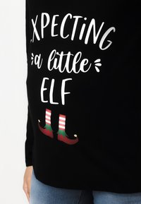 Black long-sleeve shirt featuring white text that reads "Expecting a little Elf," with graphic elf shoes at the bottom in red and green.