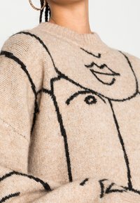 Beige stickad tröja med en svart abstrakt design av ett ansikte, rund halsringning och strukturerad tyg med synliga sömnadsdetaljer.