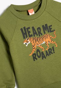 Grüner Sweatshirt mit geripptem Halsausschnitt und Bündchen. Mit einem Tiger-Graphic in Orange und Schwarz sowie "HEAR ME ROAR!" in fetter schwarzer Schrift.
