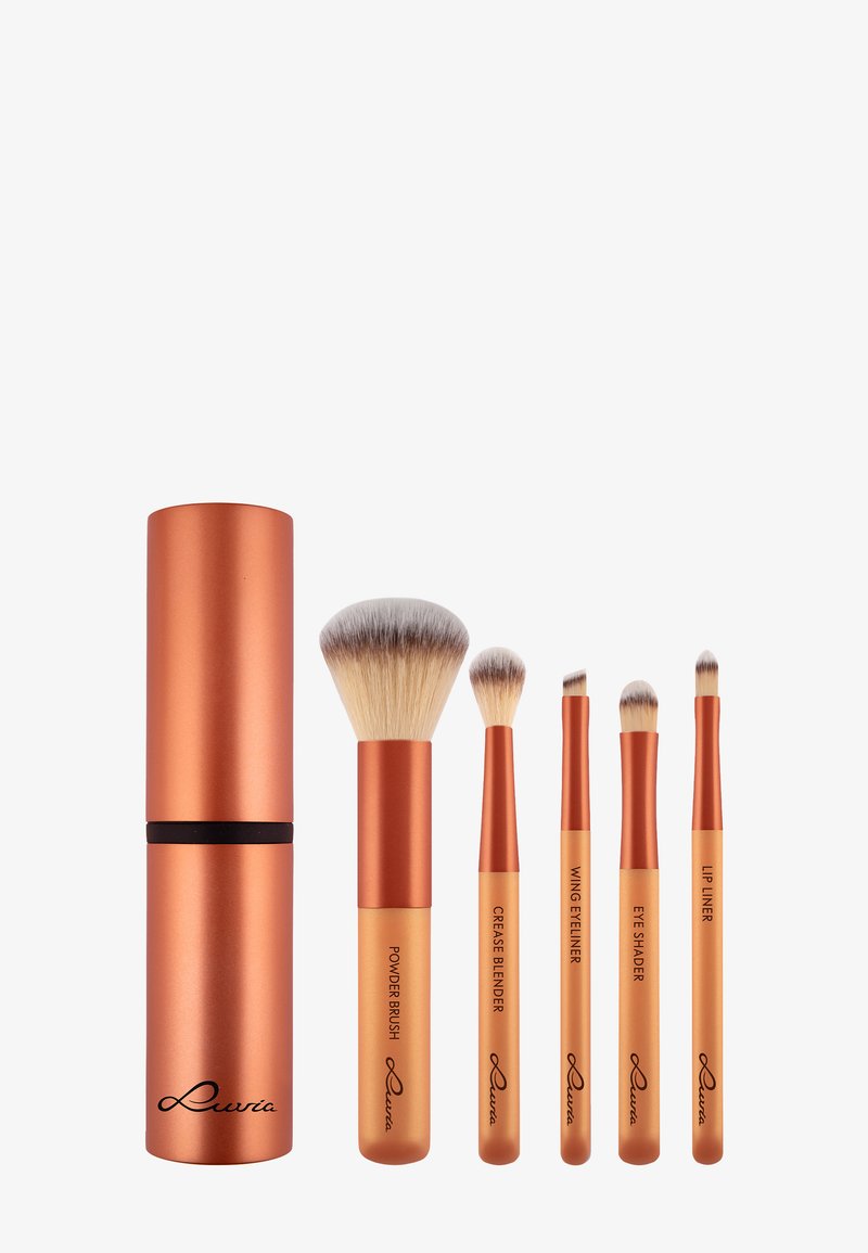 Vijf make-upkwasten in goud en koper met labels: poederkwast, crease blender, wing eyeliner, oogschaduwkwast, lip liner en een bijpassend koperen etui.