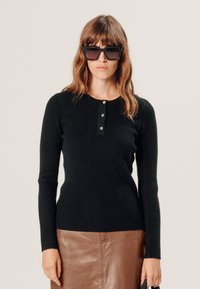 Zwarte geribbelde trui met lange mouwen en ronde halslijn, voorzien van een knoopsluiting met drie knopen. Gecombineerd met een bruine leren rok. Gladde textuur.