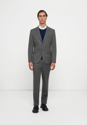 Gray suit with a structured blazer, two buttons, and notch lapels. Worn over a navy sweater with a white collared shirt. Black shoes.