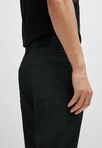 Pantalons en denim noir avec une coupe slim, arborant une étiquette de ceinture mate, des poches arrière et un discret détail texturé. Vue de profil montrant la coupe.