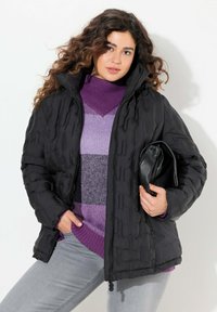 Svart vadderad jacka med texturerad yta, utrustad med hög krage och dragkedjestängning. Buren över en lila tröja och håller en svart väska.