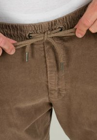 Brown corduroy joggers with an elastic waistband, featuring a drawstring closure and metallic aglets. The fabric has a ribbed texture.