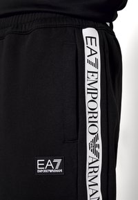 Svarta joggingbyxor med en vit sidstripe och "EA7 EMPORIO ARMANI"-logotyp, tillverkade av ett mjukt tyg med elastisk midja och logopatch.