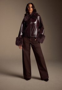 Giacca in ecopelle bordeaux con collo e polsini pelosi, abbinata a pantaloni larghi marrone scuro. Texture liscia e design casual.