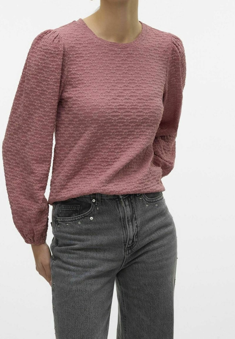 Femme portant un chemisier texturé rose poudré à manches longues et un jean noir délavé taille haute sur fond uni.