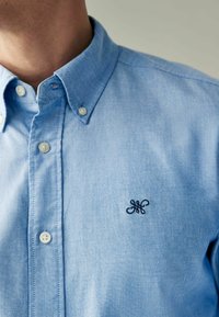 Camisa de botones azul claro hecha de algodón, con un cuello, botones blancos y un logotipo bordado oscuro en el pecho.
