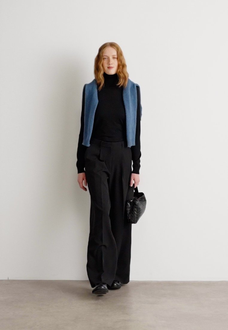 Gilet bleu coupé par-dessus un col roulé noir, assorti à un pantalon noir à jambes larges. Porte un petit sac à main noir texturé et des chaussures noires.