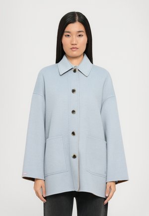 Cappotto oversize blu chiaro con colletto a punta, cinque bottoni neri e due tasche frontali. Texture morbida con dettagli di cucitura visibili.
