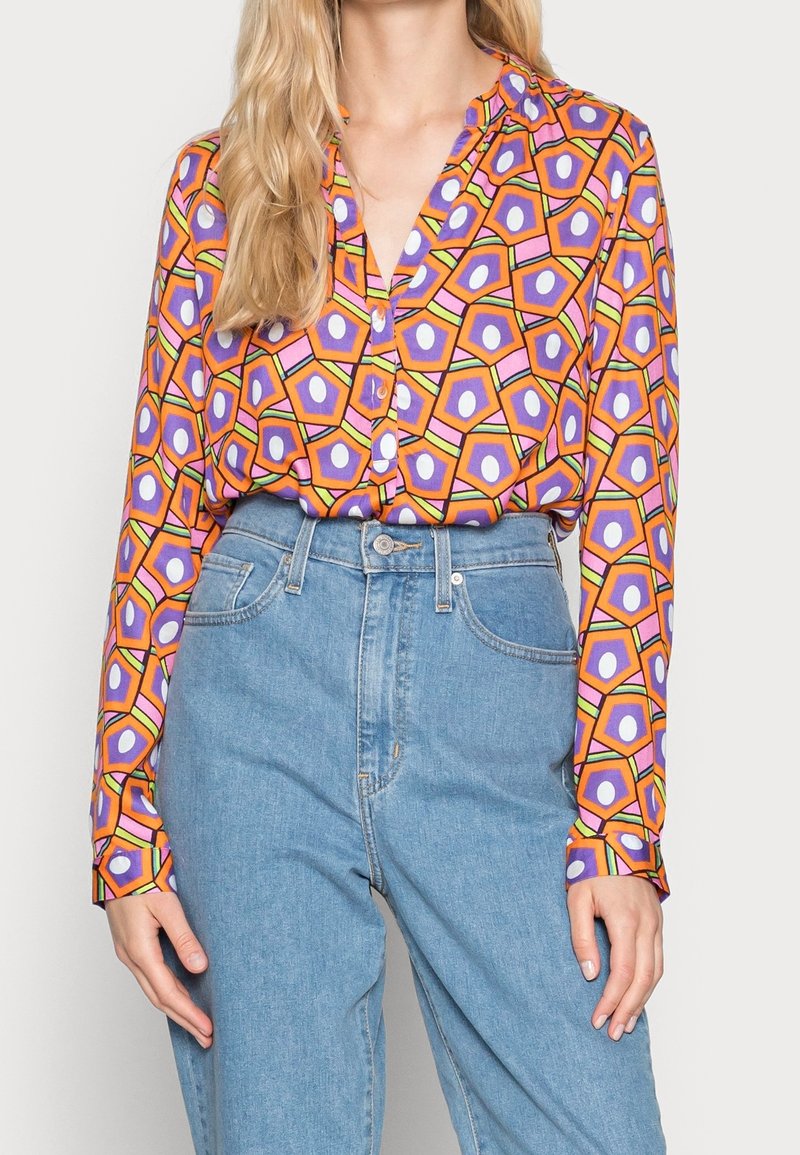 Chemise à manches longues colorée avec un motif géométrique de hexagones en orange, violet et rose sur un tissu léger. Fermeture boutonnée à l'avant.