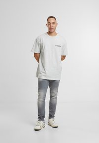 Camiseta de algodón gris con mangas cortas, que presenta un pequeño logo en el frontal. Combinada con jeans gris claros y zapatillas blancas.