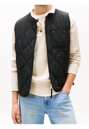 Homme portant une veste matelassée noire sur un pull henley crème et un jean bleu clair avec une main dans la poche sur fond blanc.