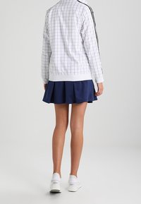 Person som bär en vit rutig jacka med svarta ränder, marinblå plisserad kjol och vita sneakers, står vänd bortåt mot en enfärgad bakgrund.