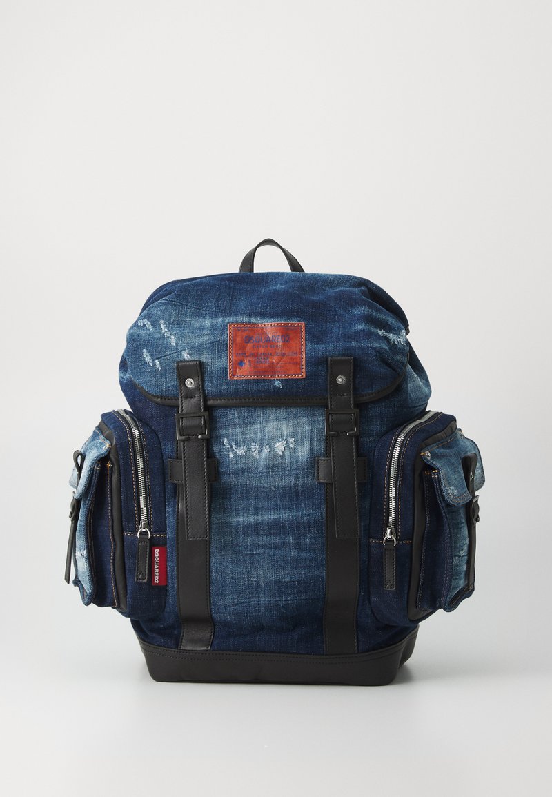 Rucsac din denim cu o formă structurată, de culoare albastru închis, cu accente din piele, buzunare frontale și fermoare metalice, având un patch din piele personalizat.
