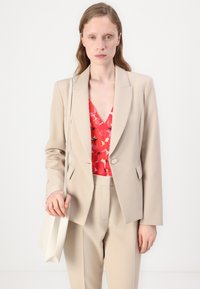 Beżowa marynarka dopasowana z wyciętymi klapami, kremowe spodnie i czerwona bluzka w kwiaty. Modelka trzyma białą torebkę, prezentując minimalistyczny design.