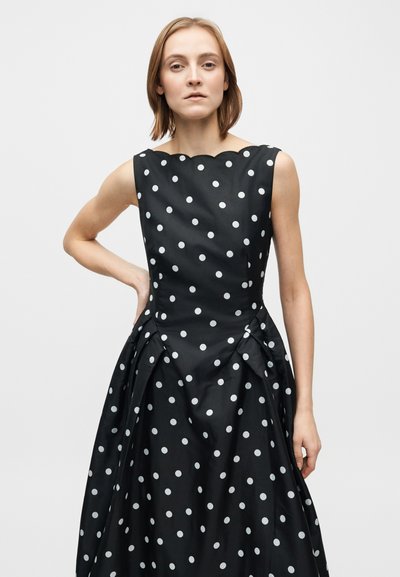 Svart klänning med vita prickar, rundad halslinning, strukturerad livdel och en flödande kjol med veck, tillverkad av ett slätt tyg.