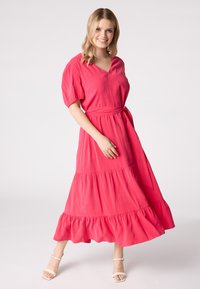 Robe maxi rose avec des manches courtes bouffantes, décolleté en V et taille ceinturée. Présente un ourlet à volants en couches et une texture lisse.