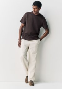 T-shirt en coton marron à manches courtes, associé à un pantalon ample beige clair et à des baskets en daim marron, sur un fond neutre.