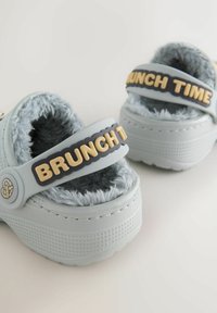 Light blue rubber sandals with a plush gray fur lining, featuring a black strap with "BRUNCH TIME" in beige letters. Textured sole.