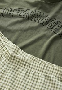 Olivgrünes T-Shirt mit dem Schriftzug "COPENHAGEN" in schwarzer Schrift über einem hellgrünen und weißen karrierten Bündchen. Weiches Baumwollmischmaterial.