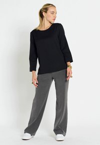 Top nero a maniche lunghe abbinato a pantaloni grigi a gamba larga; il materiale sembra liscio; accentuato da sneakers bianche e anelli audaci.