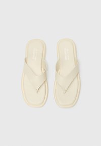 Infradito vegan beige chiaro realizzati in materiale morbido con un design a strap incrociato, suola piatta e branding minimalista sulla soletta.