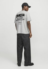 Camiseta gris de manga corta con texto gráfico negro y diseño de estrella en la parte de atrás. Combinada con pantalones de mezclilla oscuros y zapatos negros.