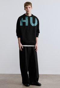 Μαύρο φούτερ με μεγάλο τιρκουάζ γράφημα "HU", σε συνδυασμό με φαρδιά μαύρα παντελόνια και μαύρα παπούτσια, τοποθετημένα σε ουδέτερο φόντο.
