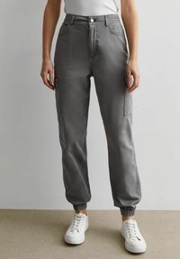 Person in grauen Cargo-Hosen mit elastischen Bündchen und weißen Sneakers, vor einem schlichten, hellen Hintergrund stehend.