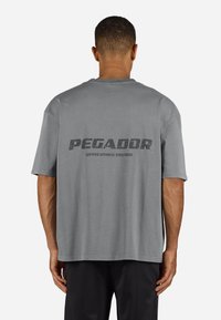 Tricou gri cu mânecă scurtă, cu gât rotund și croială supradimensionată. Are textul "PEGADOR" și branding suplimentar pe spate. Material din bumbac.