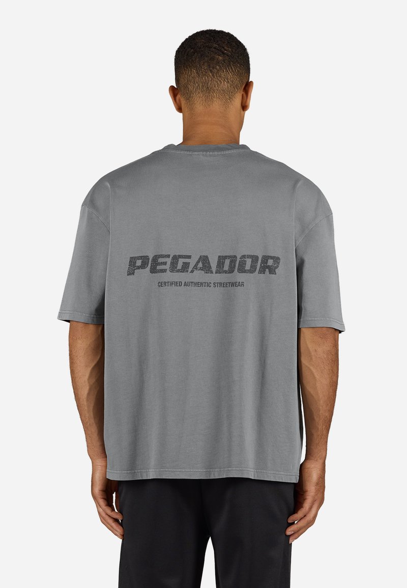 Tricou gri cu mânecă scurtă, cu gât rotund și croială supradimensionată. Are textul "PEGADOR" și branding suplimentar pe spate. Material din bumbac.