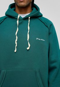Groene hoodie met een trekkoord, beige versieringen, een voorzak en 'Young Atelier' geborduurd in het wit op de borst. Katoenen materiaal.