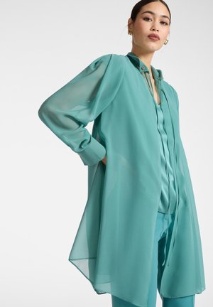 Vrouw draagt een doorschijnende teal blouse met lange mouwen over een bijpassende teal top en broek, staand met één hand in de zak tegen een effen achtergrond.