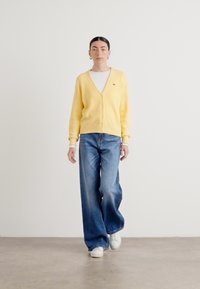 Světle žlutý cardigan s výstřihem do V a zapínáním na knoflíky, spárovaný s širokými modrými džíny a bílými teniskami. Hladká textura, uvolněný střih.