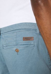 Chinos de algodón azules con un cinturón tejido de color azul claro, que presenta un parche de marca de cuero marrón y un bolsillo trasero con botón. Textura suave.