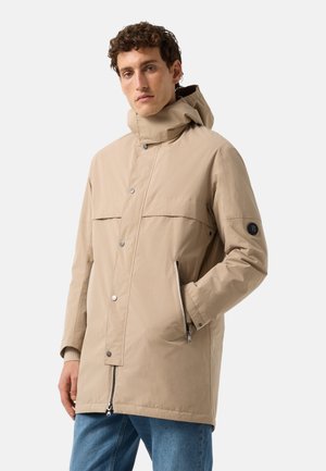 Mand iført en beige hættejakke med knapper foran og lynlåslommer, kombineret med blå jeans, stående mod en hvid baggrund.