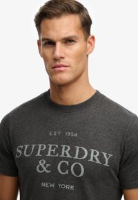 Graues Baumwoll-T-Shirt mit vorderseitigem Textmotiv "SUPERDRY & CO" und Gründungsjahr 1954. Rundhalsausschnitt und kurze Ärmel.