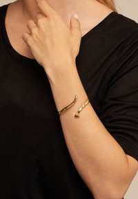 Primer plano de una persona con una camiseta negra que lleva una pulsera abierta de oro en la muñeca, con una mano tocándose el cuello.