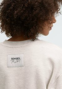 Light gray sweatshirt in soft fabric with a ribbed neckline. Features a rectangular label with the text "SENSES The Label" in black cursive.
