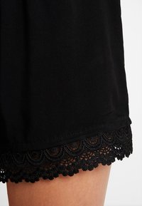 Vêtement noir avec une bordure en dentelle texturée présentant un motif en bords ondulés et de petites accents arrondis sur les bords.