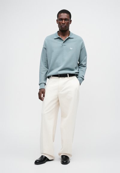 Ljusblå långärmad pikétröja med krage och broderad logotyp, tillsammans med krämfärgade vida byxor och svarta loafers.