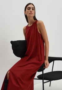 Rotes ärmelloses Kleid mit lockerer Passform, weicher Textur und seitlichen Schlitzen; kombiniert mit einer schwarzen geflochtenen Handtasche, sitzend auf einem schwarzen Stuhl.