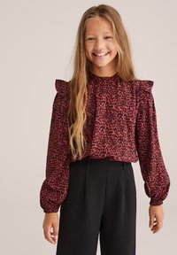 Raudona ir juoda leopardų rašto bluzonė su pūstomis rankovėmis ir raukšlėmis ant pečių, derinama su juodomis aukštai pastatytomis kelnėmis.