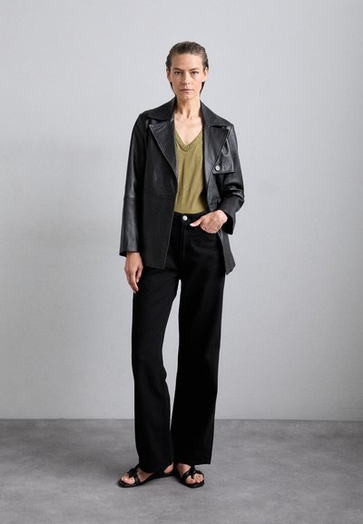 Black leather jacket with a collar, green V-neck shirt, black wide-leg pants, and black sandals, standing against a neutral background.