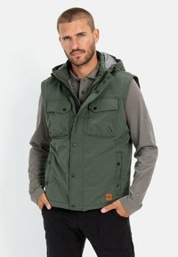 Grüne gepolsterte Weste mit Kapuze, ausgestattet mit vier Fronttaschen, Druckknopfverschluss und einem braunen Logopatch am Saum. Getragen über einem grauen Langarmshirt.